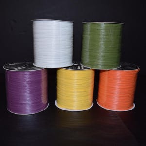 May include: Six spools of ribbon in various colors, including white, green, purple, yellow, and orange. The spools are arranged in a row on a black surface.