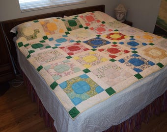 Puede incluir: Una colcha blanca con un diseño de patchwork que presenta cuadrados de varios patrones florales y geométricos en tonos de azul, verde, amarillo, rojo y rosa. La colcha está hecha con una variedad de telas y se muestra en una cama con una sábana blanca y una falda de cama a rayas.