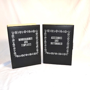 Könnte beinhalten: Zwei schwarze, rechteckige Aufbewahrungsboxen mit hellen Kanten. Jede Box hat einen weißen Blumenrahmen um den zentrierten Text. Die linke Box trägt die Aufschrift MONOGRAMMER AND TEMPLATES, die rechte ACCESSORIES AND BUTTONHOLER.