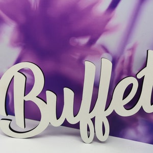 Könnte beinhalten: Ein weißes Holzschild mit dem Wort "Buffet" in kursivschrift. Das Schild befindet sich auf einem lila und weißen Hintergrund.
