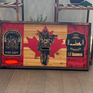 May include: Wooden wall art depicting a Canadian flag with a firefighter silhouette and fire department emblems. The artwork is framed in dark wood, with red and natural wood tones.