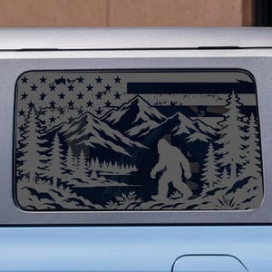 May include: A dark gray decal applied to the rear side window of a light blue vehicle. The decal features a silhouette of a Bigfoot creature walking through a forest with mountains in the background, overlaid with a stylized American flag pattern.
