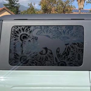Peut inclure: Un SUV vert clair avec une vitre noire ornée d'un motif décoratif de taureau entouré de tournesols. Le véhicule est garé à l'extérieur par une journée ensoleillée.