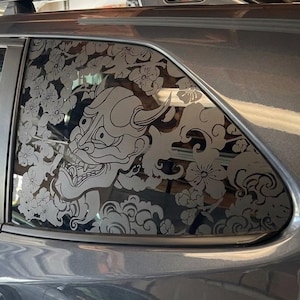 May include: A dark gray vehicle features a large, intricate white vinyl decal applied to the rear passenger side window. The design depicts a traditional Japanese Hannya mask surrounded by stylized cherry blossoms and swirling cloud patterns.