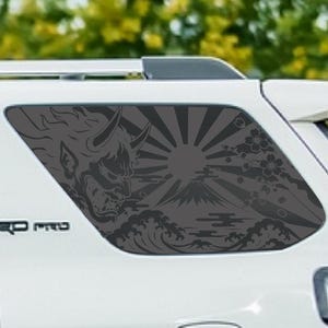 May include: A dark gray vinyl decal applied to the rear side window of a white vehicle. The decal features a detailed illustration of a Japanese Hannya mask, a rising sun with radiating beams, Mount Fuji, stylized ocean waves, and cherry blossoms.