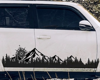 May include: A white SUV with a black roof rack and a black and white mountain and compass decal on the side. The vehicle is parked in a snowy area.
