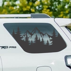 May include: A dark gray vinyl decal applied to the rear side window of a white vehicle. The decal features a silhouette design of a mountain range surrounded by a dense forest of pine trees. The text TRD PRO is visible on the vehicle body.