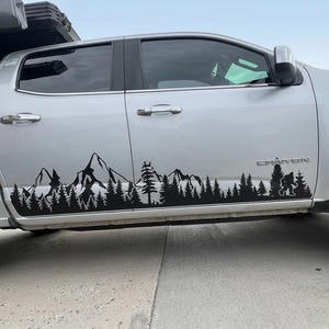 May include: A silver pickup truck featuring a black vinyl decal applied to the lower side panels. The decal displays a continuous landscape graphic consisting of jagged mountain peaks, a dense forest of pine trees, and a silhouette of Bigfoot.
