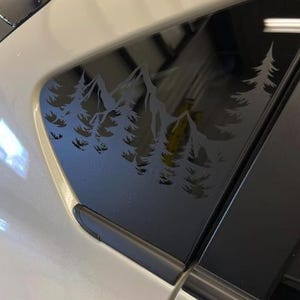 K&ouml;nnte beinhalten: Ein matt-dunkelgrauer Vinyl-Aufkleber auf einer Autoscheibe, der eine detaillierte Silhouette eines Gebirges umgeben von einem dichten Kiefernwald zeigt. Das Design ist ausgeschnitten, sodass das Glas durch den Zwischenraum sichtbar ist.
