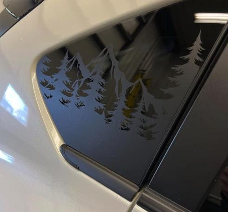 K&ouml;nnte beinhalten: Ein matt-dunkelgrauer Vinyl-Aufkleber auf einer Autoscheibe, der eine detaillierte Silhouette eines Gebirges umgeben von einem dichten Kiefernwald zeigt. Das Design ist ausgeschnitten, sodass das Glas durch den Zwischenraum sichtbar ist.