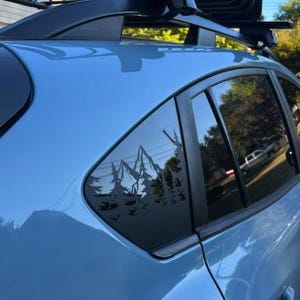 K&ouml;nnte beinhalten: Ein hellblaues Fahrzeug ist mit einem schwarzen Vinyl-Aufkleber am hinteren Seitenfenster versehen. Der Aufkleber zeigt die detaillierte Silhouette eines Gebirges, das von einem dichten Kiefernwald umgeben ist und sich vom Glas abhebt.
