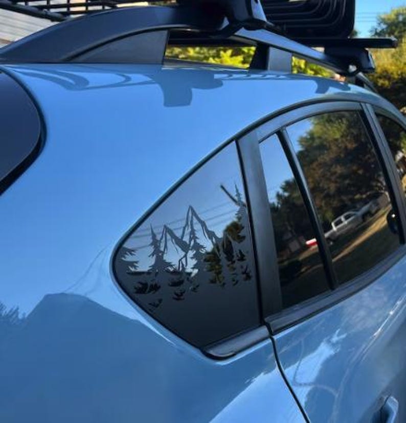 K&ouml;nnte beinhalten: Ein hellblaues Fahrzeug ist mit einem schwarzen Vinyl-Aufkleber am hinteren Seitenfenster versehen. Der Aufkleber zeigt die detaillierte Silhouette eines Gebirges, das von einem dichten Kiefernwald umgeben ist und sich vom Glas abhebt.