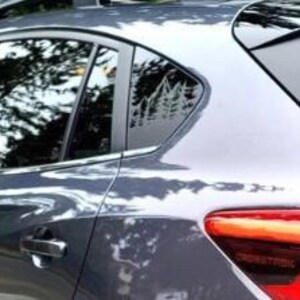 K&ouml;nnte beinhalten: Ein graues Auto ist mit einem wei&szlig;en Vinyl-Aufkleber am hinteren Seitenfenster versehen. Der Aufkleber zeigt eine detaillierte Grafik eines Gebirgszuges mit Tannen an der Basis, die eine von der Natur inspirierte Silhouette auf dem Glas bildet.