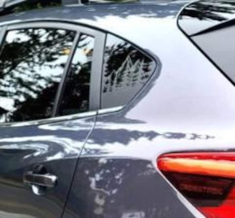 K&ouml;nnte beinhalten: Ein graues Auto ist mit einem wei&szlig;en Vinyl-Aufkleber am hinteren Seitenfenster versehen. Der Aufkleber zeigt eine detaillierte Grafik eines Gebirgszuges mit Tannen an der Basis, die eine von der Natur inspirierte Silhouette auf dem Glas bildet.