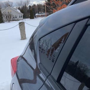 K&ouml;nnte beinhalten: Ein dunkelgraues Fahrzeug ist mit einem wei&szlig;en Vinyl-Aufkleber auf dem hinteren Seitenfenster versehen. Der Aufkleber zeigt ein stilisiertes Gebirge mit Nadelb&auml;umen im Vordergrund, das einen scharfen Kontrast zur dunklen Fensterscheibe bildet.