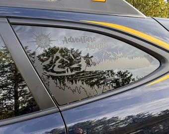 May include: A matte grey vinyl decal applied to a car window featuring a mountain range, a dense forest, a compass rose, and a person paddleboarding with a dog. The text Adventure Awaits is printed in a cursive script above the mountain peaks.