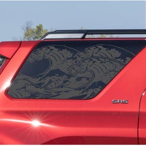 May include: A dark grey vinyl decal featuring a stylised ocean wave design applied to the rear side window of a bright red vehicle. The graphic depicts a large, cresting wave with intricate foam details, covering the entire glass surface area.