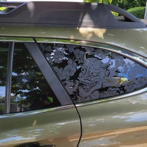 May include: A dark olive green vehicle features a black vinyl decal applied to the rear quarter window. The decal displays a detailed black line art illustration of a Japanese Hannya mask surrounded by stylized cherry blossoms and swirling clouds.