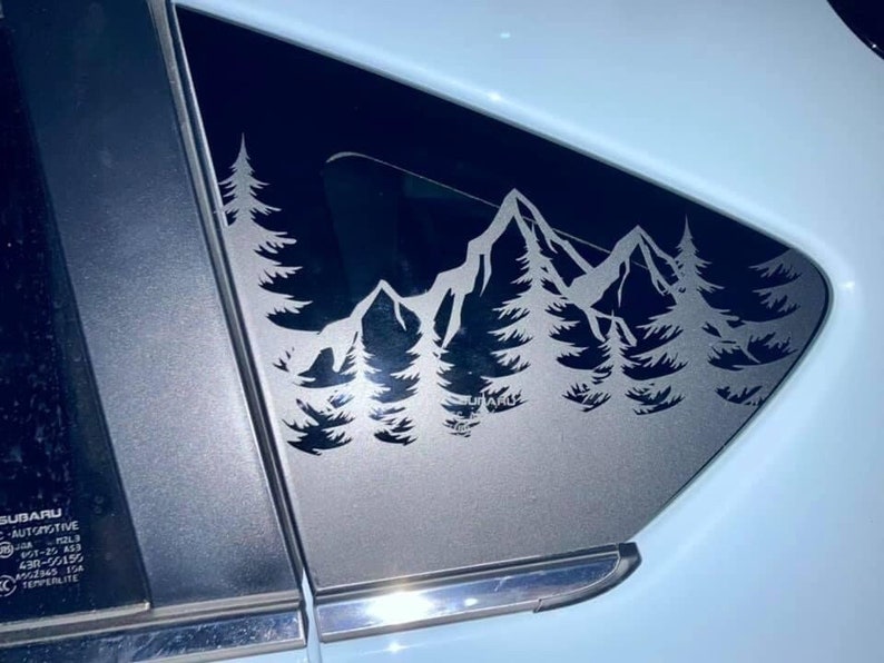 K&ouml;nnte beinhalten: Ein silberfarbener Vinyl-Aufkleber auf einer Autoscheibe, der ein detailliertes Landschaftsmotiv mit gezackten Berggipfeln und einem dichten Nadelwald zeigt. Das Design ist pr&auml;zise ausgeschnitten und bildet einen Kontrast zum dunklen Glas.