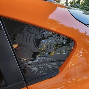 May include: A black vinyl decal applied to the triangular rear side window of an orange vehicle. The decal features a detailed graphic design of a large, stylized ocean wave with intricate foam patterns and swirling water currents.