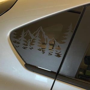 K&ouml;nnte beinhalten: Ein mattschwarzer Vinyl-Aufkleber auf einer Autoscheibe, der eine gestanzte Silhouette eines Gebirges mit Kiefern im Vordergrund zeigt. Das Design ist einfarbig und haftet auf der Glasoberfl&auml;che, wobei die Landschaft als Negativraum erscheint.
