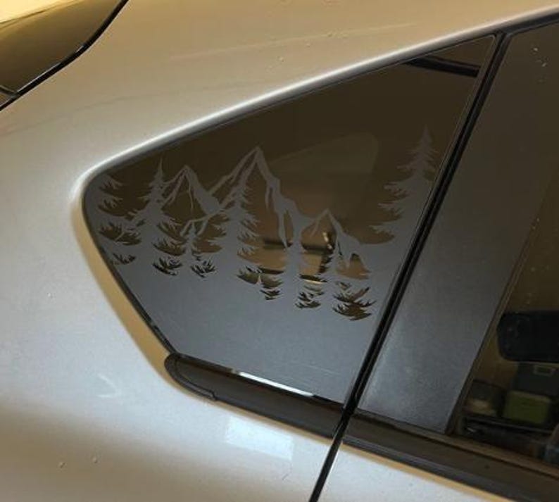 K&ouml;nnte beinhalten: Ein mattschwarzer Vinyl-Aufkleber auf einer Autoscheibe, der eine gestanzte Silhouette eines Gebirges mit Kiefern im Vordergrund zeigt. Das Design ist einfarbig und haftet auf der Glasoberfl&auml;che, wobei die Landschaft als Negativraum erscheint.