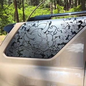 May include: A tan SUV with a decorative window graphic featuring a detailed illustration of a horned oni mask surrounded by floral patterns. The design is in shades of gray and black.