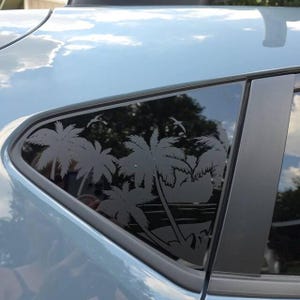 May include: A dark gray vinyl decal applied to a car window, featuring a tropical beach scene with multiple palm trees, a setting sun on the horizon, and two birds flying in the sky. The design is cut out to show the dark tint of the glass.