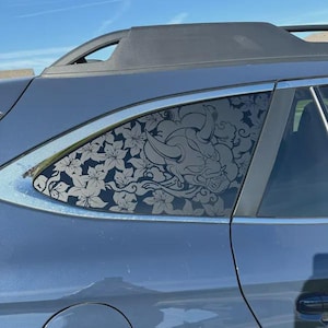 May include: A dark gray car window decal featuring a detailed line art illustration of a horned demon mask surrounded by stylized floral patterns and swirling cloud motifs, applied to the rear side window of a dark blue vehicle.