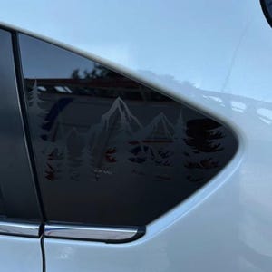 K&ouml;nnte beinhalten: Ein wei&szlig;es Auto hat ein dunkel get&ouml;ntes hinteres Seitenfenster, das mit einem wei&szlig;en Vinyl-Aufkleber verziert ist. Der Aufkleber zeigt eine detaillierte Landschaftsgrafik mit gezackten Berggipfeln und einem dichten Kiefernwald, wobei das Wort Subaru sichtbar ist.