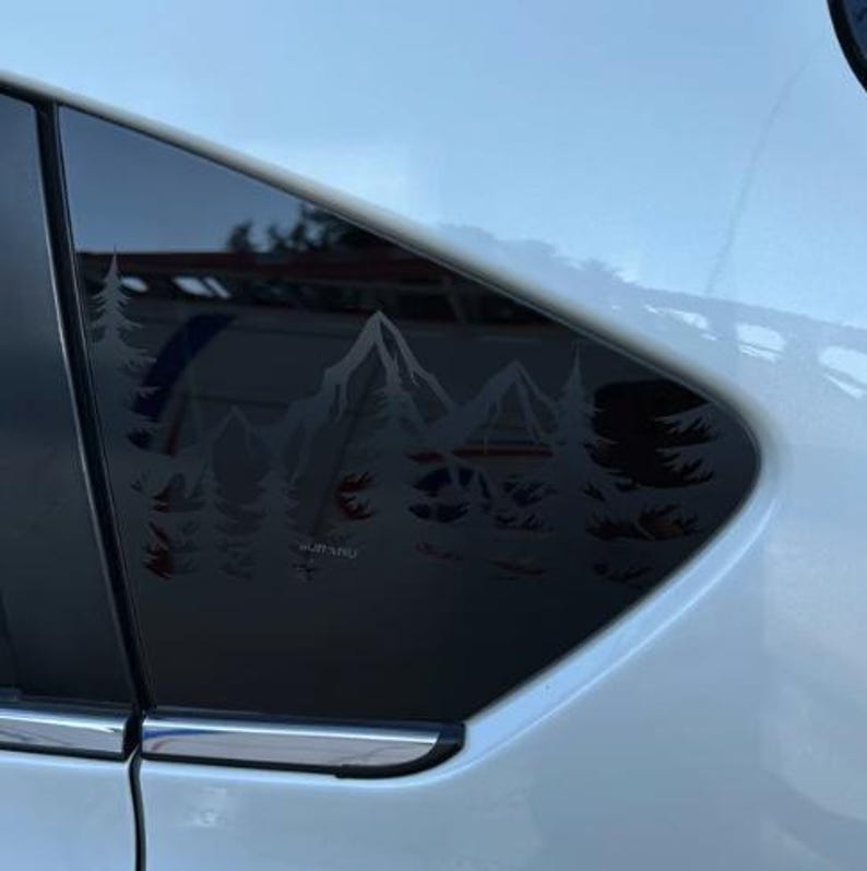 K&ouml;nnte beinhalten: Ein wei&szlig;es Auto hat ein dunkel get&ouml;ntes hinteres Seitenfenster, das mit einem wei&szlig;en Vinyl-Aufkleber verziert ist. Der Aufkleber zeigt eine detaillierte Landschaftsgrafik mit gezackten Berggipfeln und einem dichten Kiefernwald, wobei das Wort Subaru sichtbar ist.