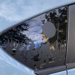 May include: A black vinyl decal applied to a car window featuring a silhouette of a Bigfoot creature walking through a dense forest of pine trees with a large full moon and several small stars positioned in the sky above the mountain peaks.