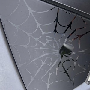 May include: A black, glossy, triangular-shaped decal featuring a spiderweb pattern. The design consists of thin, curved lines radiating from a central point, creating a web effect against the solid black surface of the adhesive material.