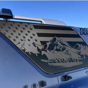 May include: A matte black decal applied to a vehicle's rear window, featuring a stylized American flag design with a mountain range and forest scene. The text "DDC" is visible on the top right. The bottom text reads "DEFAULT MATTE BLACK MATERIAL".