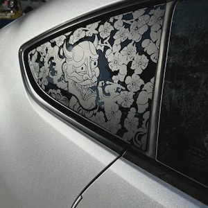 May include: A silver car window decal featuring a white Hannya mask design surrounded by detailed cherry blossom flowers and swirling cloud patterns. The graphic is applied to the triangular rear quarter window of a metallic silver vehicle.