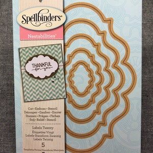May include: A pack of five Spellbinders Nestabilities dies for crafting. The dies are shaped like ornate frames with a scalloped edge. The packaging is brown and white with the text "Spellbinders Nestabilities" and "Thankful for you".