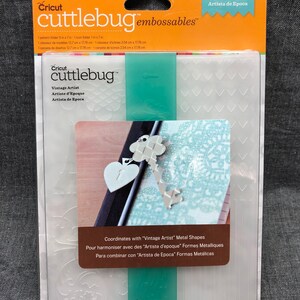 May include: A clear plastic sheet with a Cricut cuttlebug embossables design. The sheet features a pattern of hearts, keys, and other decorative elements. The text "Vintage Artist" is printed on the sheet. The sheet is packaged in a clear plastic sleeve with a yellow and orange background.