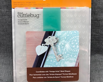 May include: A clear plastic sheet with a Cricut cuttlebug embossables design. The sheet has a pattern of hearts, keys, and other decorative elements. The text "Vintage Artist" is printed on the sheet. The sheet is packaged in a clear plastic sleeve with a yellow and orange background.