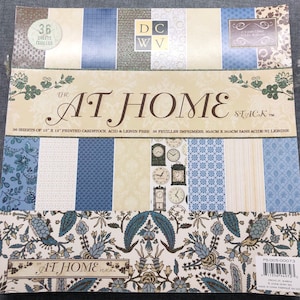 May include: A pack of 36 sheets of 12x12 inch acid and lignin free printed cardstock featuring a blue and white floral pattern with a clock theme. The pack is labeled "The At Home Stack" and includes the text "36 sheets of 12" x 12" printed cardstock acid & lignin free 36 feuilles imprimées, 30.5cm x 30.5cm sans acide ni lignine".