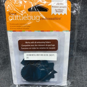 May include: A package of Cricut Cuttlebug embossable metal shapes. The package is orange and white and features a picture of the shapes. The shapes are silver and include a variety of designs, including a circle, a star, and a heart. The package also includes the text "CB EMB METAL GIRLY GIRL SILVER (2002197) IP:3".