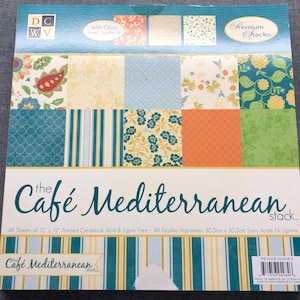May include: A pack of 48 sheets of 12x12 inch printed cardstock with a Mediterranean theme. The paper is acid and lignin free. The pack features a variety of patterns including floral, striped, and geometric designs in shades of blue, green, yellow, and orange. The text "The Cafe Mediterranean stack" is printed on the front of the pack.