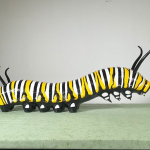 May include: A black, white and yellow striped caterpillar sculpture with a long, thin body and two long, thin antennae. The caterpillar is made of a smooth, hard material and has a glossy finish.