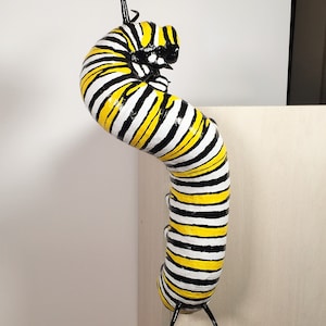 May include: A black and yellow striped caterpillar sculpture with black antennae. The caterpillar is posed in a curled position, as if it is about to transform into a butterfly.