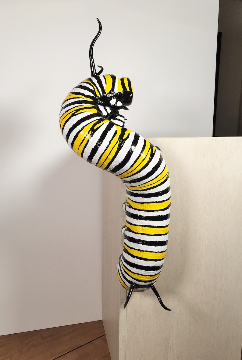 May include: A black and yellow striped caterpillar sculpture with black antennae. The caterpillar is posed in a curled position, as if it is about to transform into a butterfly.
