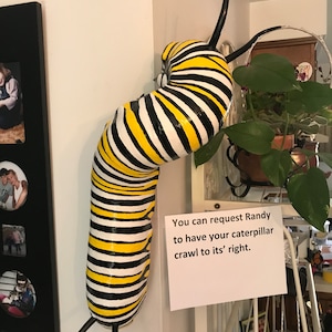 May include: A black, yellow, and white striped caterpillar sculpture hanging on a wall. The sculpture has two long black antennae and is curled up in a spiral shape. A white sign with black text reads "You can request Randy to have your caterpillar crawl to its' right."
