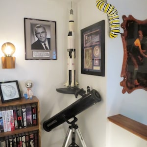 May include: A black telescope on a tripod, a model rocket, a framed photo of a man, a framed photo with a US flag, a wooden shelf with books, a small figurine, and a yellow and black caterpillar on the wall.