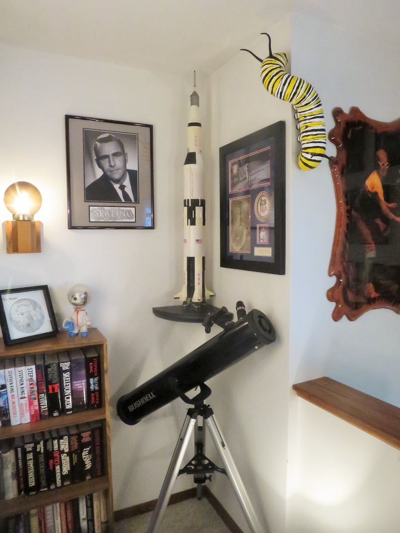 May include: A black telescope on a tripod, a model rocket, a framed photo of a man, a framed photo with a US flag, a wooden shelf with books, a small figurine, and a yellow and black caterpillar on the wall.