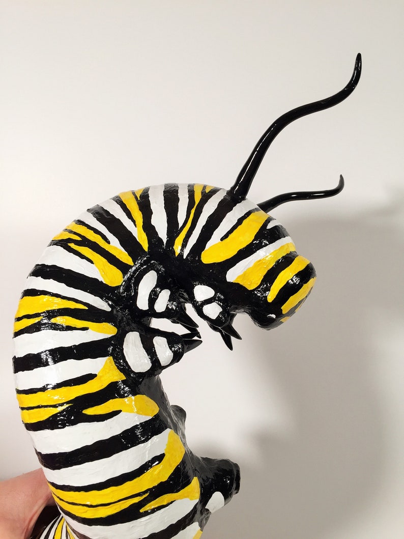 May include: A black and yellow striped caterpillar sculpture with black antennae. The caterpillar is curled up in a C shape.