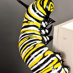 May include: A black and yellow striped caterpillar sculpture with a white background. The caterpillar is curled up and appears to be climbing a surface.