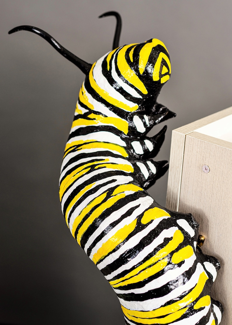May include: A black and yellow striped caterpillar sculpture with a white background. The caterpillar is curled up and appears to be climbing a surface.